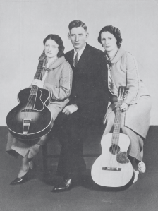 black and white phot of a daughter, father, and mother sitting on a bench. The women are in wool coats and holding guitars. The father is in the middle with a black suit